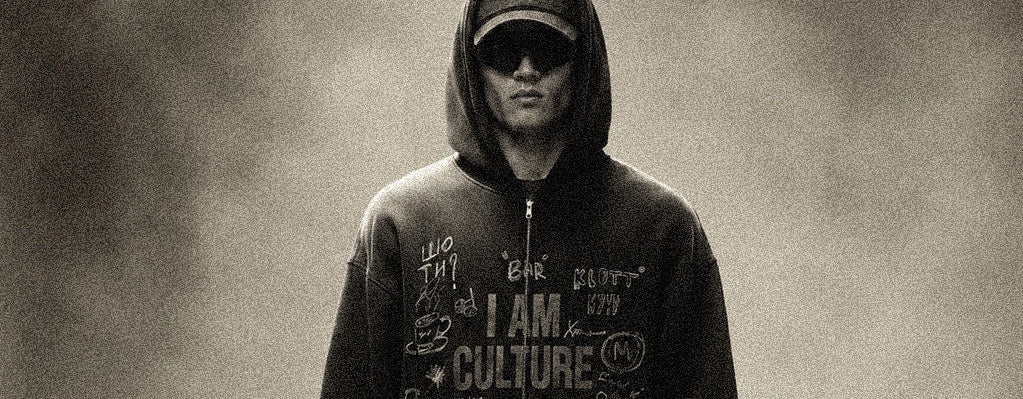 A person stands in a smoky studio setting wearing a dark graphic zip-up hoodie with the words 'I AM CULTURE' across the front, dark pants, and sneakers, while holding a dark bag.